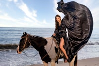 a woman in a black dress riding a horse on the beach
